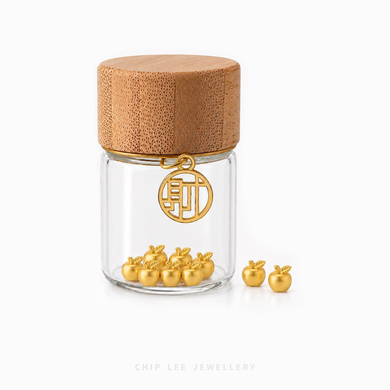 A clear glass jar with a round bamboo lid and a gold hanging charm, shown on a white background with several small gold apple-shaped jewelry pieces placed in front and inside the jar. The brand name 'CHIP LEE JEWELLERY' appears faintly at the bottom.