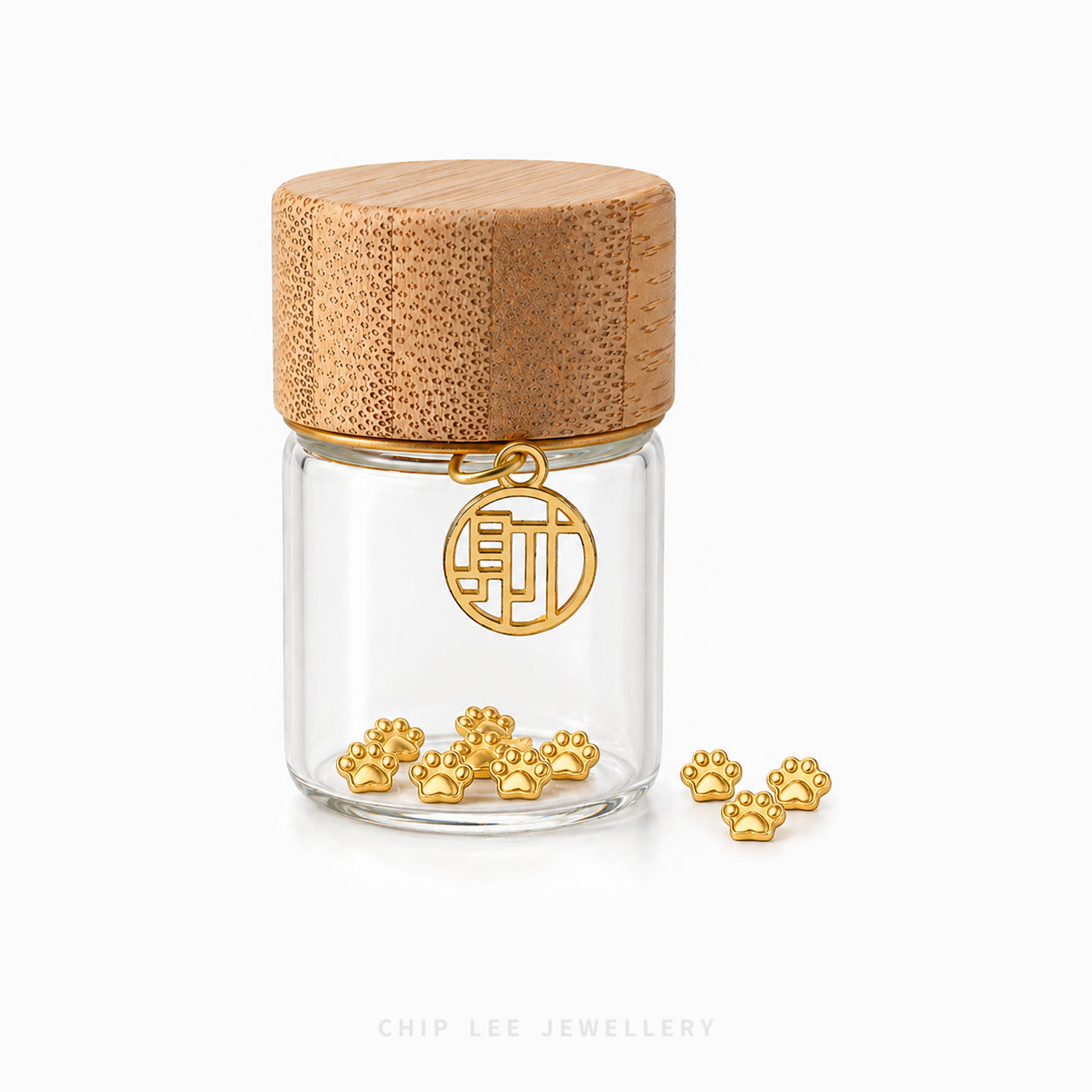 A clear glass jar with a light brown bamboo lid, featuring a gold-toned circular hanging charm and several small gold paw-shaped charms scattered at the base, on a white background.