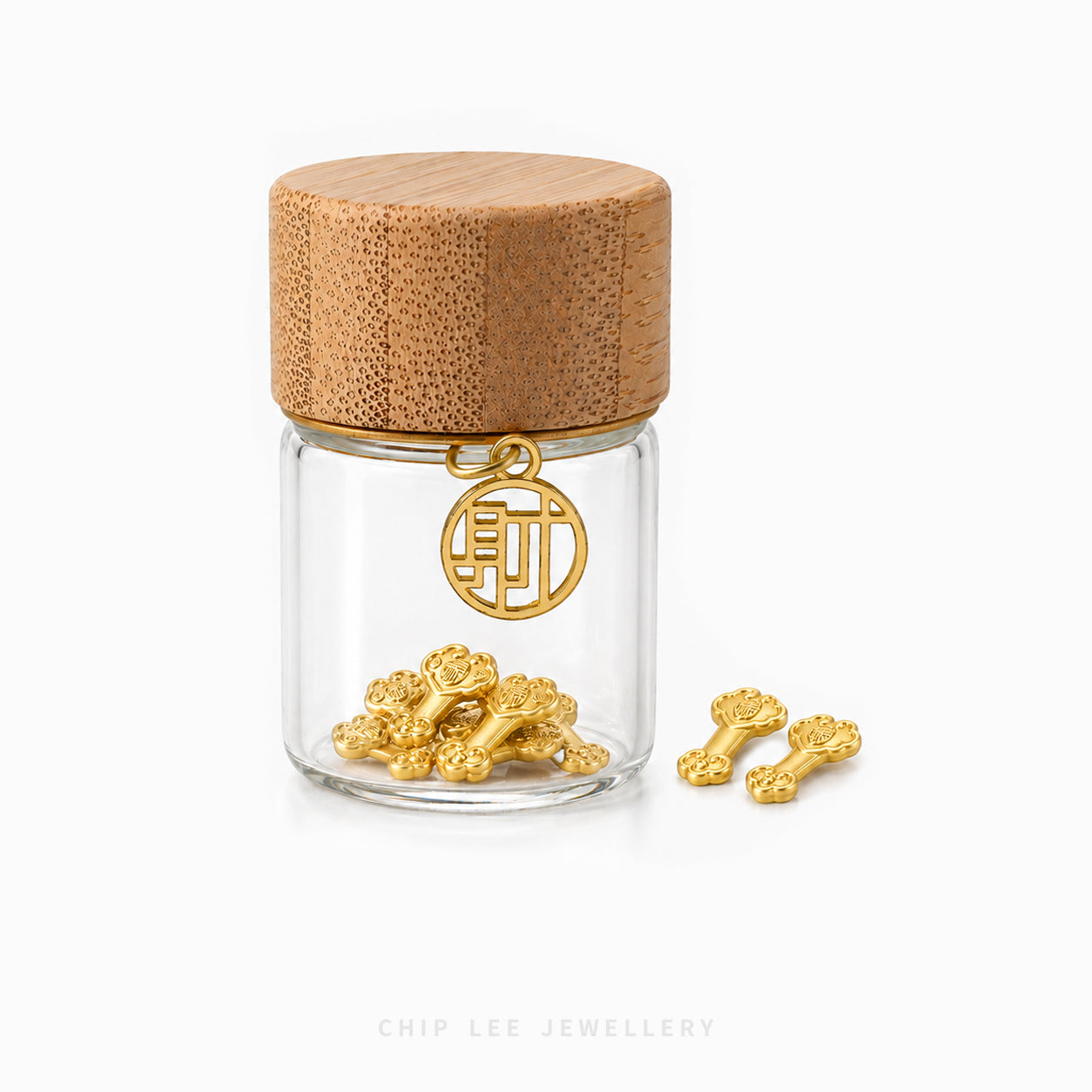 A small clear glass jar with a light wood lid holds several gold flower-shaped charms, with one charm hanging from the jar and two more placed beside it on a white background. The text 'CHIP LEE JEWELLERY' appears faintly at the bottom.