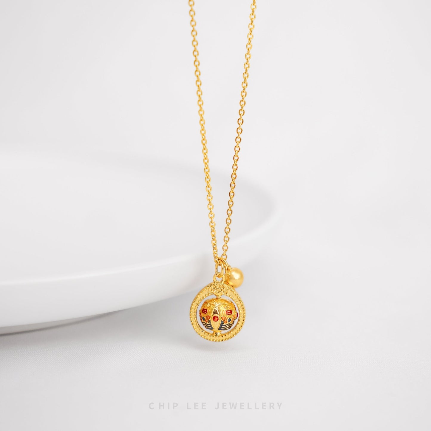 A gold necklace with a circular spinning charm pendant in the center, featuring intricate designs, and a small gold bell on the end of the chain.