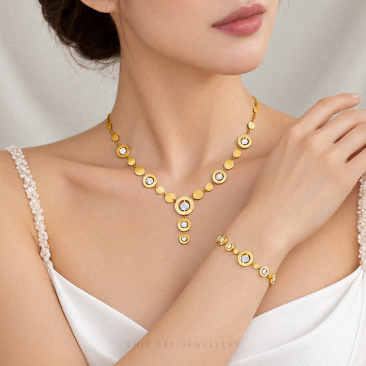 A close-up lifestyle shot of a model wearing a gold necklace and matching bracelet from Chip Lee Jewellery. The jewelry features a gold-plated circular disc design with clear rhinestones.