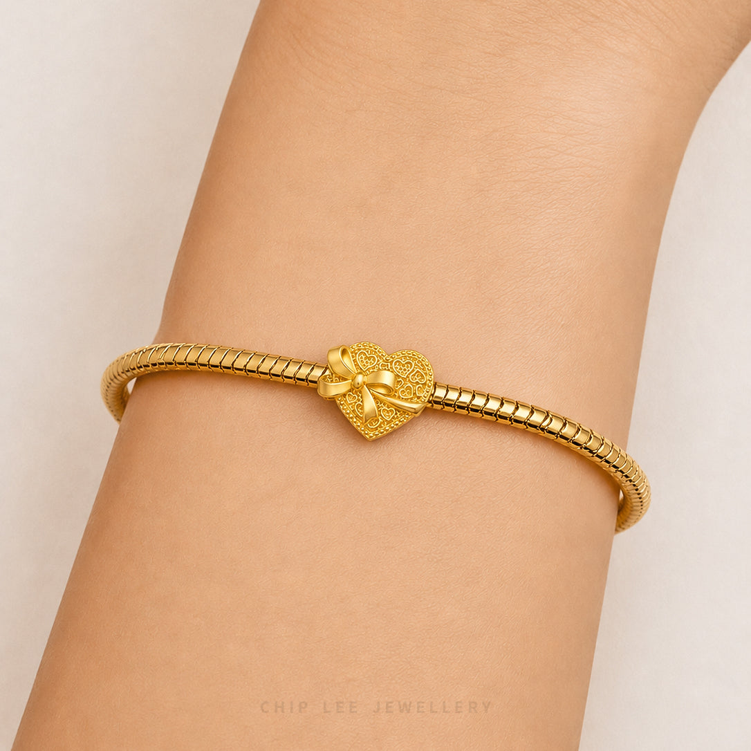 A gold snake-braided bracelet with a textured heart charm featuring a small bow, shown worn on a person's wrist against a light neutral background.