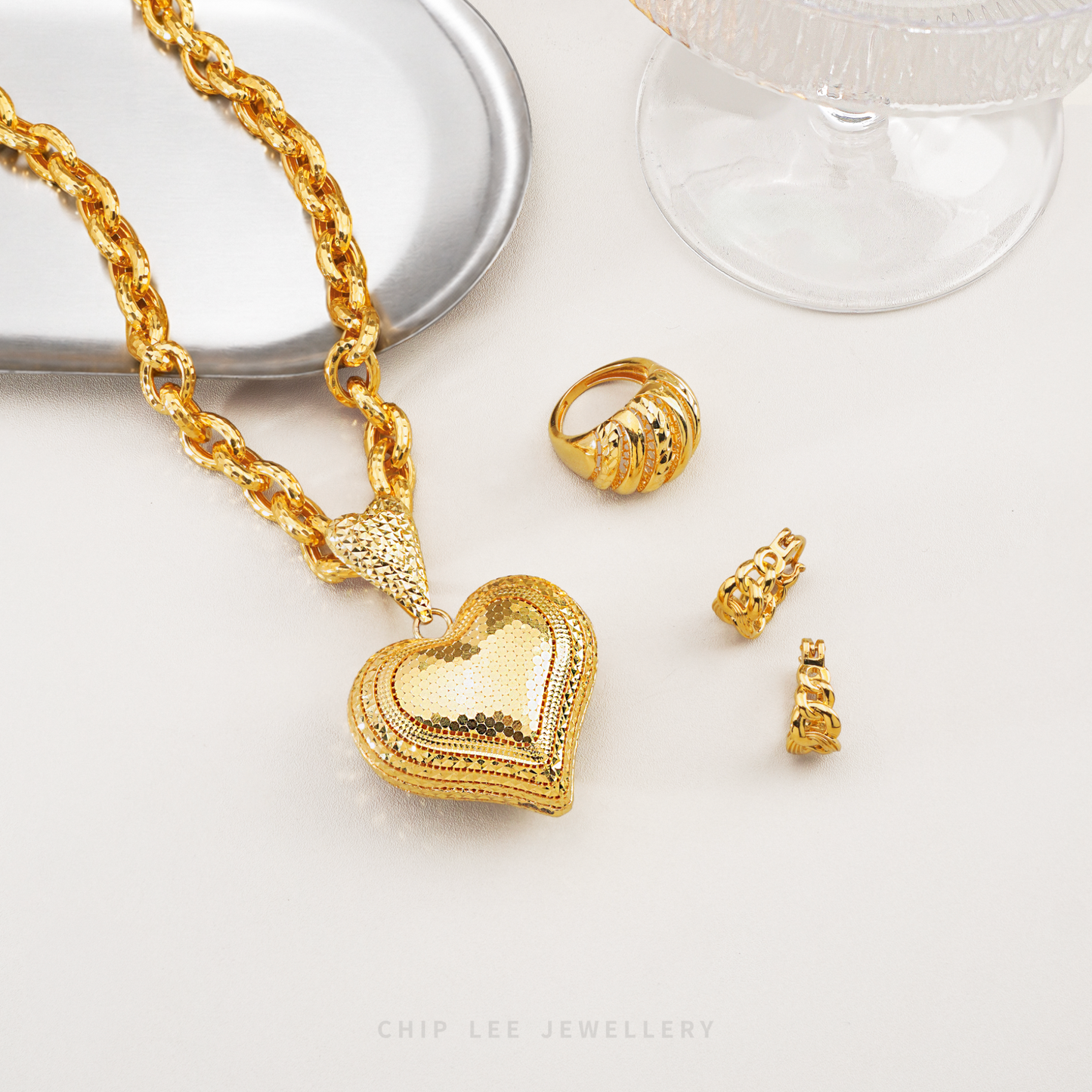 Gold heart-shaped necklace, ring, and earrings on a white surface with 'Chip Lee Jewellery' branding.
