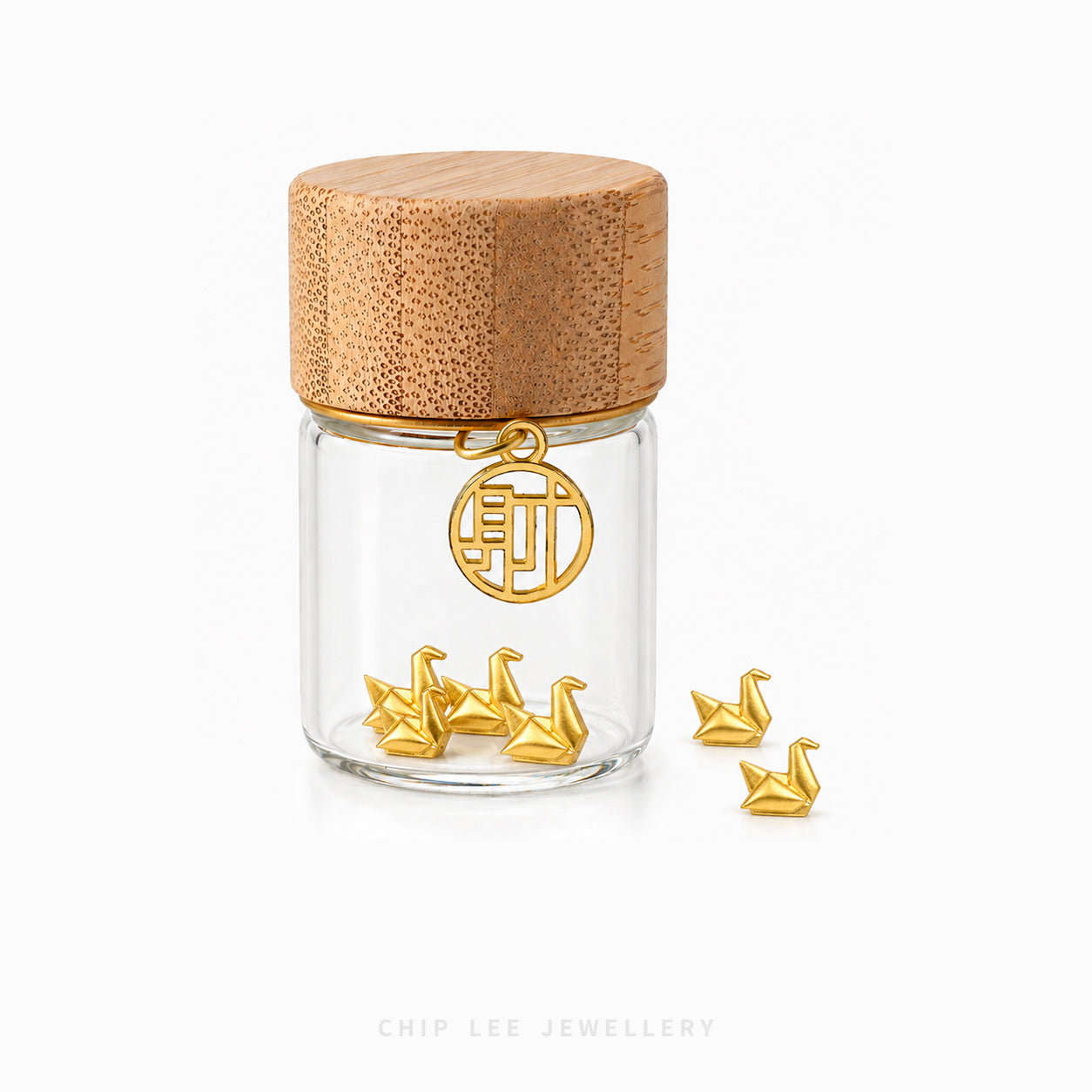 A clear glass jar with a light wood lid, featuring a gold-tone hanging charm with a Chinese character and several small gold swan-shaped jewelry pieces inside and beside the jar, on a white background. Chip Lee Jewellery branding is visible at the bottom.
