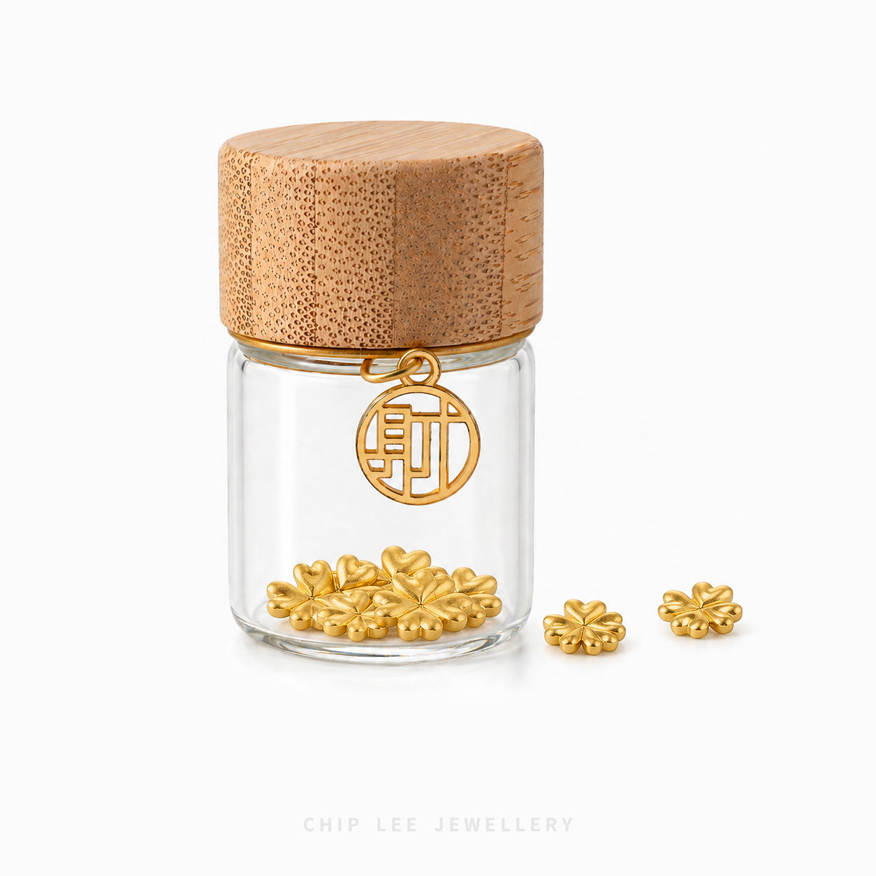 A small clear glass jar with a light wood lid, containing small gold flower-shaped jewelry pieces, with two matching gold pieces placed beside it on a white background. A circular gold charm hangs from the jar.