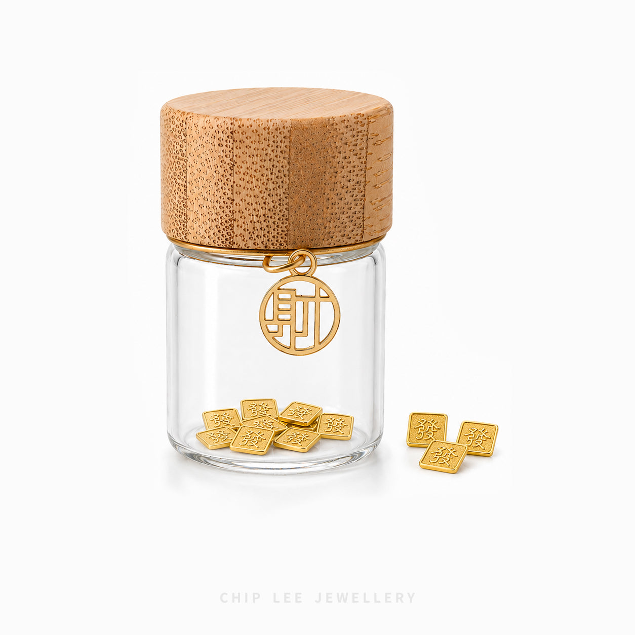 A clear glass jar with a textured light brown bamboo lid, featuring a gold circular pendant with Chinese characters and several small gold square charms placed inside and beside the jar on a white background. The brand name 'CHIP LEE JEWELLERY' is printed at the bottom.