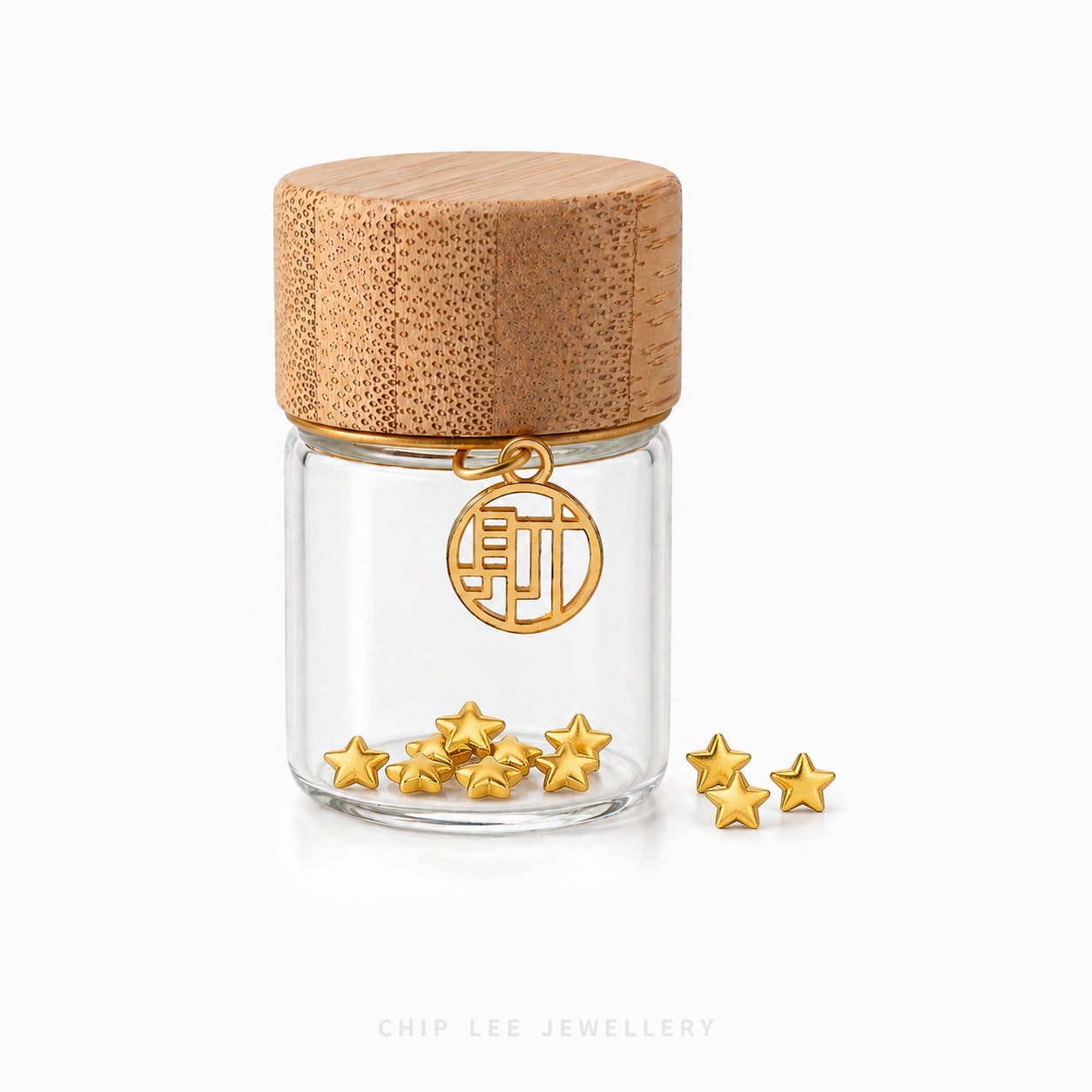 A small clear glass jar with a light wood lid and a gold-tone hanging charm, shown on a white background with several small gold star-shaped jewelry pieces beside it. The brand name 'CHIP LEE JEWELLERY' appears faintly at the bottom.