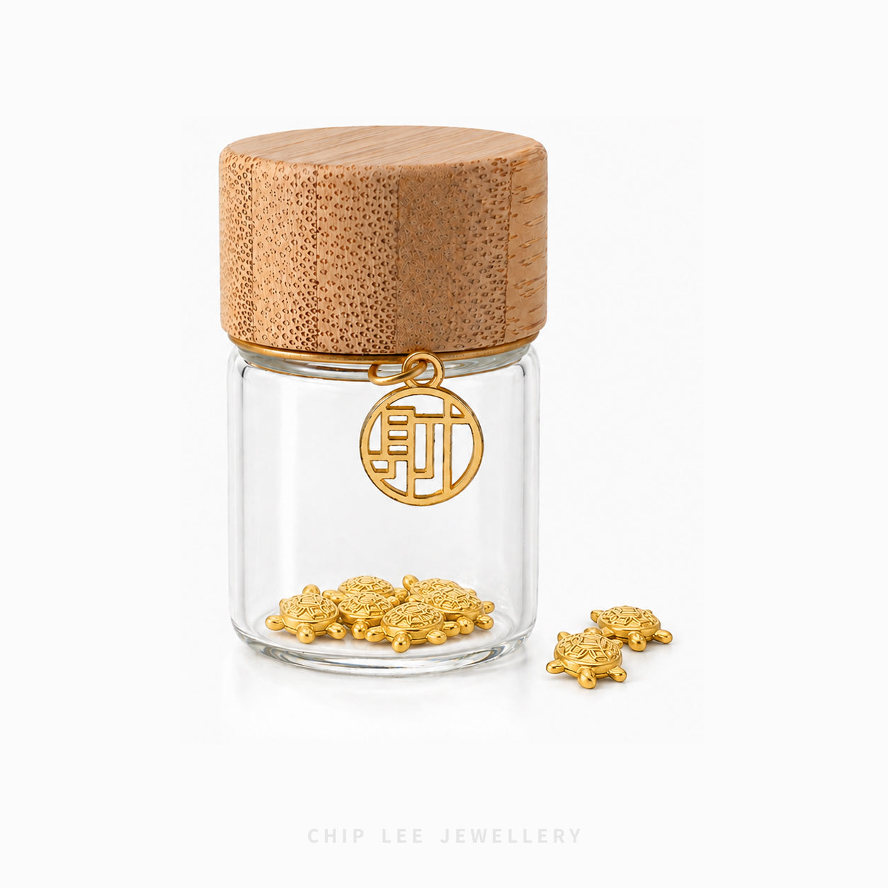 A small clear glass jar with a light wood lid and a gold-tone circular charm featuring a Chinese character, shown on a white background with small gold charms placed beside it. The brand name 'CHIP LEE JEWELLERY' appears faintly at the bottom.