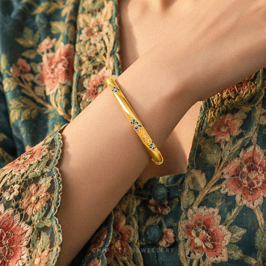 A close-up of a person's wrist wearing a gold bangle bracelet with blue floral details, paired with a green floral lace top.