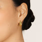 Close-up of a model wearing a small gold hoop earring with polished and textured floral details, her dark hair styled in a low bun against a cream background.