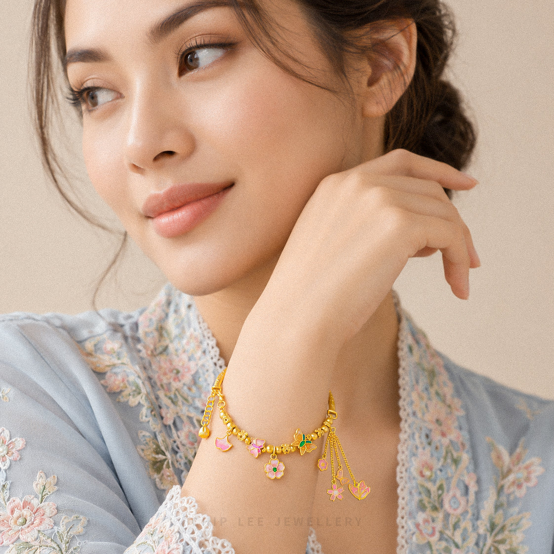 A woman wearing a delicate gold chain bracelet with small dangling charms, including pink hearts, green beads, and a flower, posed against a soft beige background.