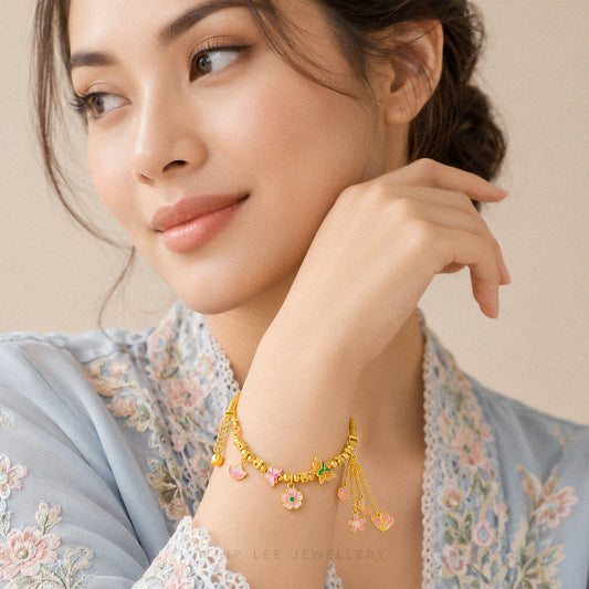 A woman wearing a delicate gold chain bracelet with small dangling charms, including pink hearts, green beads, and a flower, posed against a soft beige background.