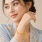 A woman wearing a delicate gold chain bracelet with small dangling charms, including pink hearts, green beads, and a flower, posed against a soft beige background.