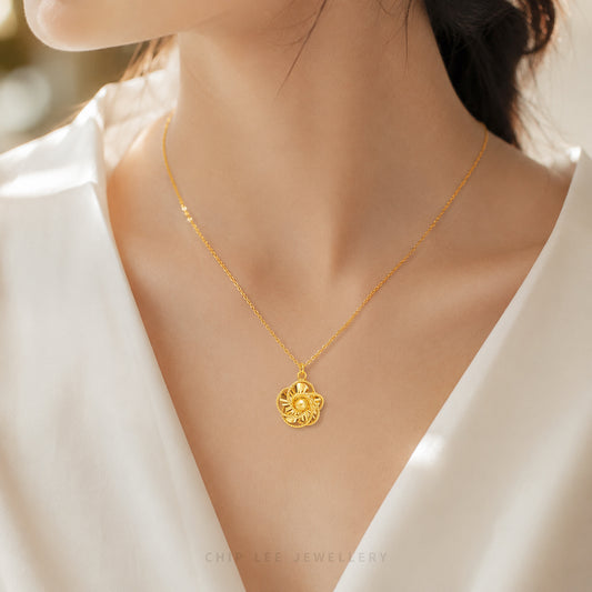 A delicate gold chain necklace with a small flower-shaped pendant worn by a model in a white V-neck top. The pendant features a sunburst design with a central circular detail.