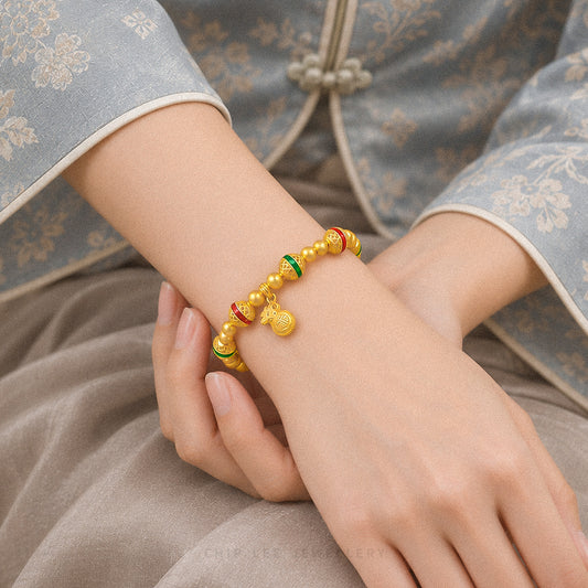 A close-up of a person's wrist wearing a gold beaded bracelet with red and green accent beads and a small hanging charm, styled with a traditional patterned blue-gray garment.