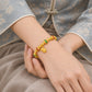 A close-up of a person's wrist wearing a gold beaded bracelet with red and green accent beads and a small hanging charm, styled with a traditional patterned blue-gray garment.