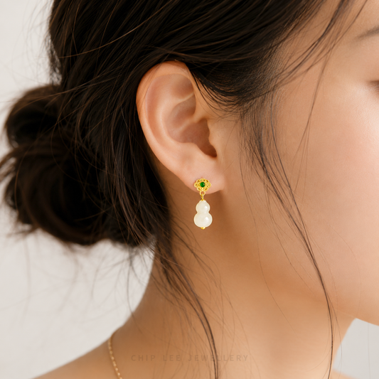 A close-up of a model's ear wearing a gold dangle earring with a small green stone and two white pearl-like drops, against a soft light gray background.
