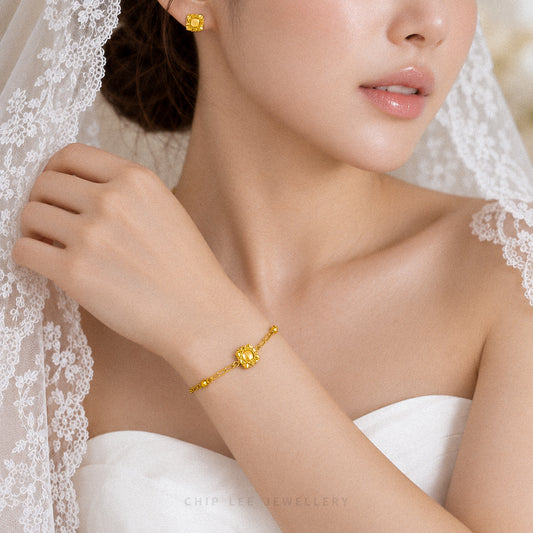 A close-up lifestyle shot of a bride wearing a delicate gold chain bracelet with a central flower-shaped gold charm. She is wearing a white lace-trimmed wedding dress and veil, with a matching gold flower stud earring visible.