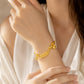 A close-up lifestyle shot of a woman wearing a delicate gold bracelet on her wrist. The bracelet features a series of small, circular textured gold medallions and a small dangling charm.