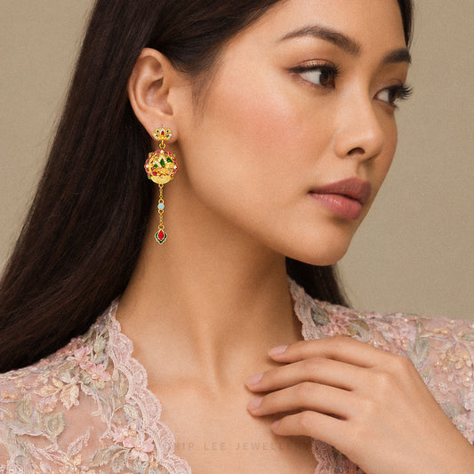 A close-up lifestyle shot of a woman wearing a gold-plated traditional Indian Kundan dangle earring adorned with red, green, and blue stones. The model is wearing a light pink lace top against a neutral beige background.