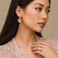 A close-up lifestyle shot of a woman wearing a gold-plated traditional Indian Kundan dangle earring adorned with red, green, and blue stones. The model is wearing a light pink lace top against a neutral beige background.
