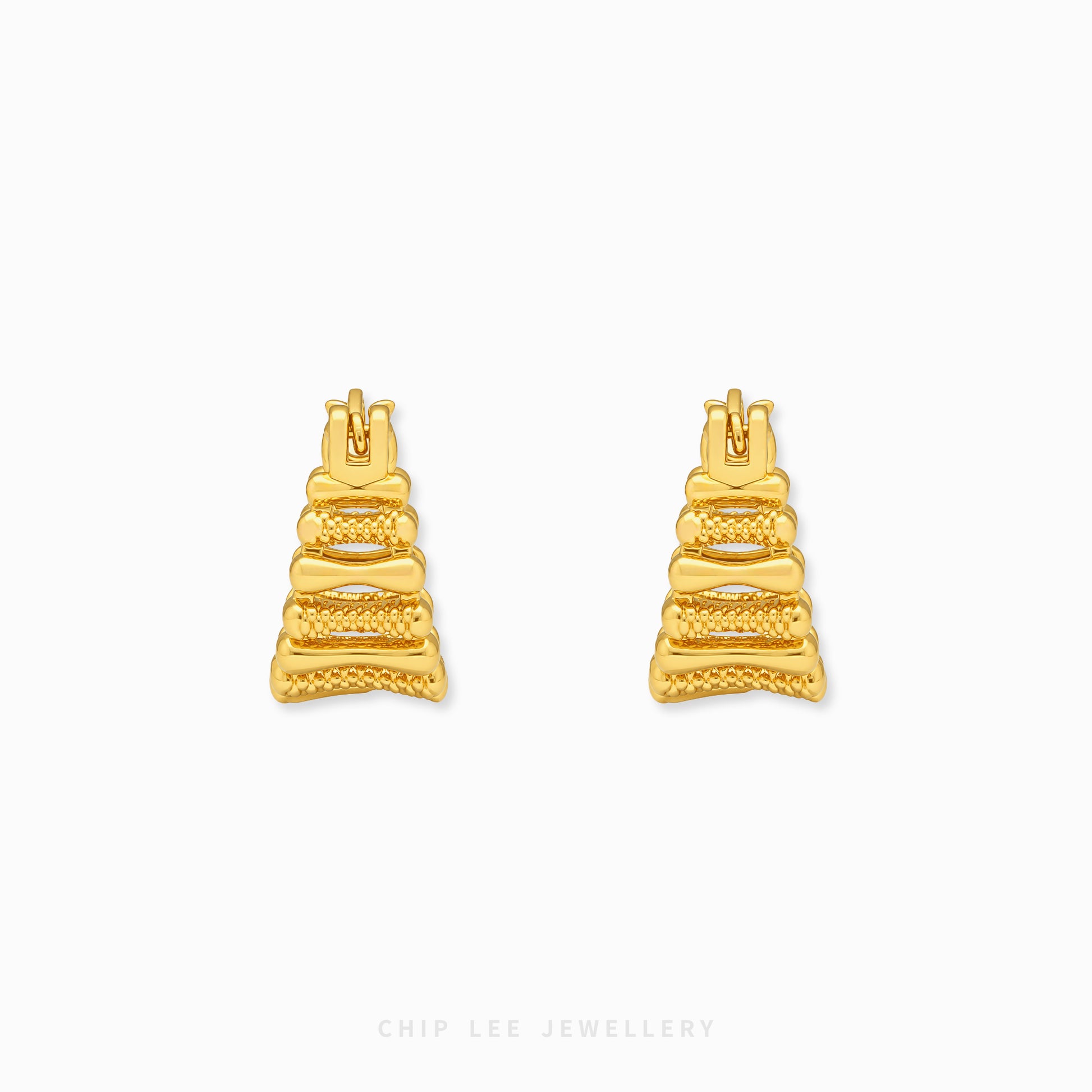 A pair of gold geometric stud earrings with a textured, tiered design, displayed on a clean white background with a subtle brand watermark at the bottom.