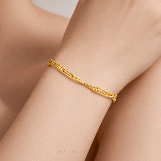 A delicate gold bangle bracelet with a twisted, textured design, shown worn on a person's wrist. The background consists of soft-focus skin tones.
