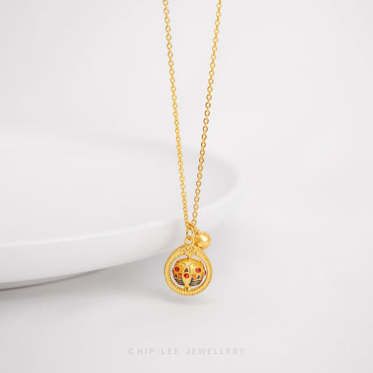 A gold necklace with a circular spinning charm pendant in the center, featuring intricate designs, and a small gold bell on the end of the chain.