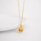 A gold necklace with a circular spinning charm pendant in the center, featuring intricate designs, and a small gold bell on the end of the chain.