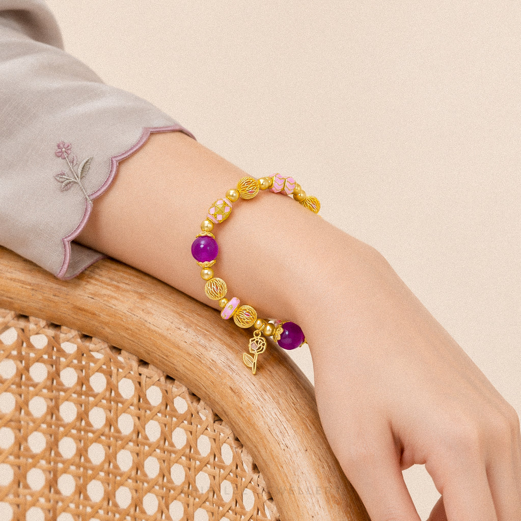 A gold beaded bracelet with purple and pink beads, a small gold leaf charm, and a tiny bird charm, shown worn on a person's wrist resting on a wooden chair.
