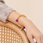 A gold beaded bracelet with purple and pink beads, a small gold leaf charm, and a tiny bird charm, shown worn on a person's wrist resting on a wooden chair.