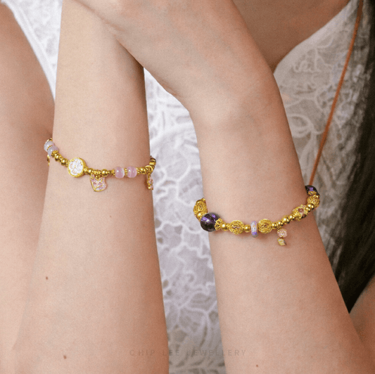 Close-up of crossed wrists wearing delicate 916 gold bead bracelets with pastel gemstone accents and symbolic charms