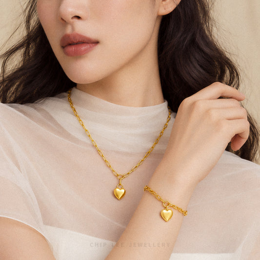 A close-up lifestyle shot of a model wearing a gold heart pendant necklace and a matching gold heart bracelet, styled with a sheer white t-shirt against a soft beige background.