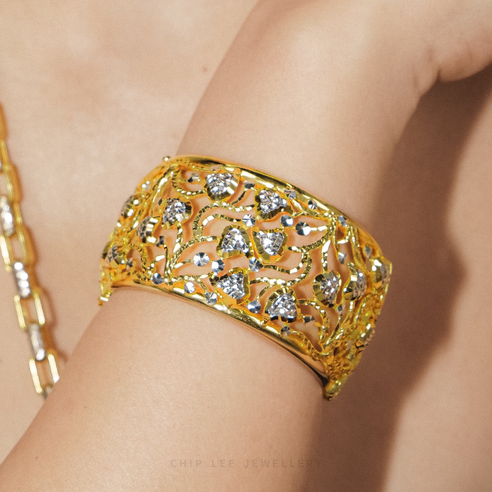 A close-up of a gold filigree cuff bracelet with clear gemstones worn on a person's wrist. The intricate openwork pattern is highlighted by warm lighting, and a matching gold chain is partially visible on the left.