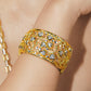 A close-up of a gold filigree cuff bracelet with clear gemstones worn on a person's wrist. The intricate openwork pattern is highlighted by warm lighting, and a matching gold chain is partially visible on the left.