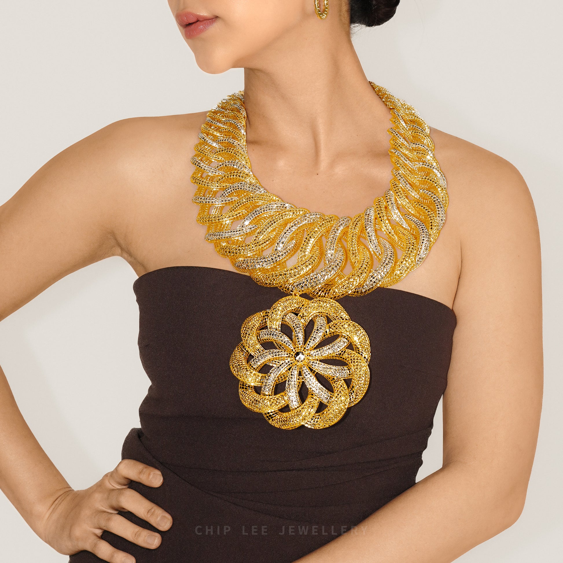 A close-up of a model wearing a dramatic chunky gold statement necklace with silver pavé accents and a matching large flower-shaped brooch, paired with a dark brown strapless dress.