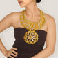 A close-up of a model wearing a dramatic chunky gold statement necklace with silver pavé accents and a matching large flower-shaped brooch, paired with a dark brown strapless dress.