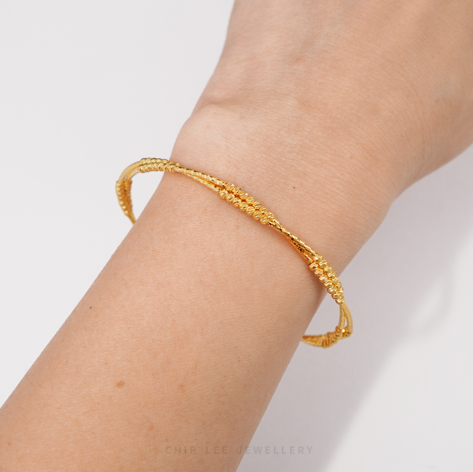 A textured gold bangle bracelet worn on a person's wrist, shown against a clean white background with a brand watermark at the bottom.