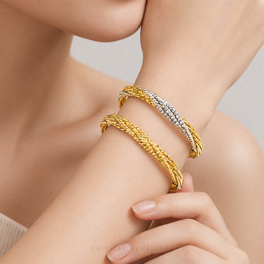 Two twisted bangle bracelets on a model's wrist, one in yellow gold and one in yellow gold with a pavé diamond-look silver inlay, against a soft neutral background.