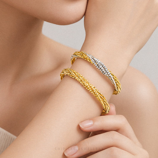 Two twisted bangle bracelets on a model's wrist, one in yellow gold and one in yellow gold with a pavé diamond-look silver inlay, against a soft neutral background.