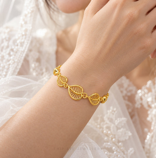 A delicate gold bracelet with three interlocking leaf motifs worn on a woman's wrist, set against a soft white lace dress background.
