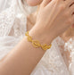 A delicate gold bracelet with three interlocking leaf motifs worn on a woman's wrist, set against a soft white lace dress background.
