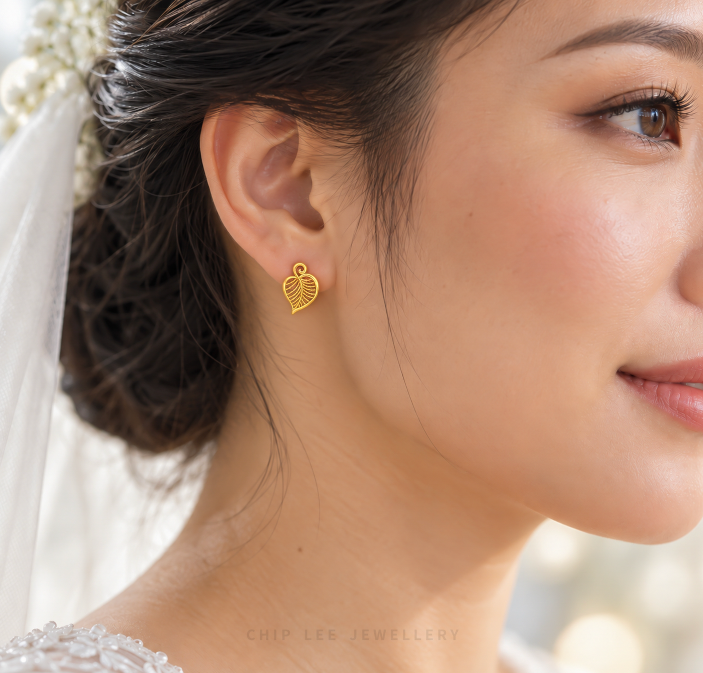A close-up of a woman's ear wearing a small gold heart-shaped stud earring with a leaf-like openwork design. She has dark hair pulled back and is wearing a white bridal veil and dress, with a soft blurred background.