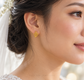 A close-up of a woman's ear wearing a small gold heart-shaped stud earring with a leaf-like openwork design. She has dark hair pulled back and is wearing a white bridal veil and dress, with a soft blurred background.