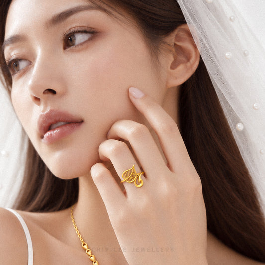 A close-up lifestyle shot of a model wearing a gold necklace and matching gold ring with a leaf motif. The jewelry features intricate filigree and textured details, presented on a model in a bridal-style white dress with a veil.