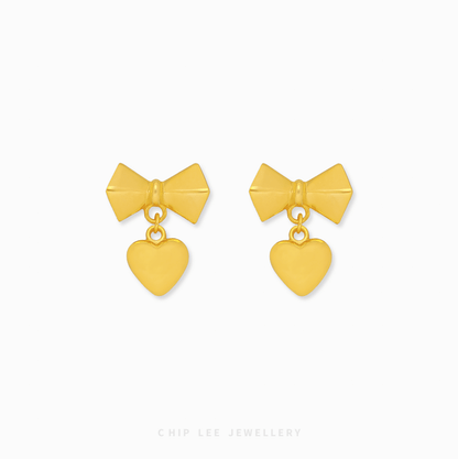 A pair of 999 pure gold bow dangling heart earrings showing the fluid movement and high polish of the 24K yellow gold on a dark background.

