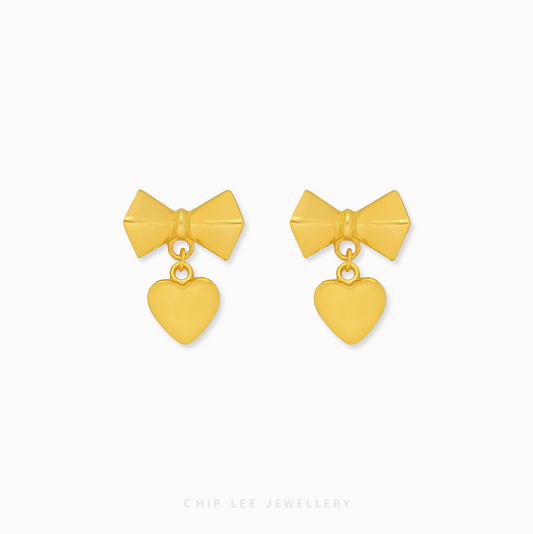 A pair of 999 pure gold bow dangling heart earrings showing the fluid movement and high polish of the 24K yellow gold on a dark background.

