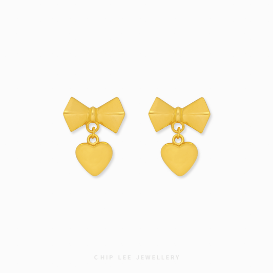 A pair of 999 pure gold bow dangling heart earrings showing the fluid movement and high polish of the 24K yellow gold on a dark background.