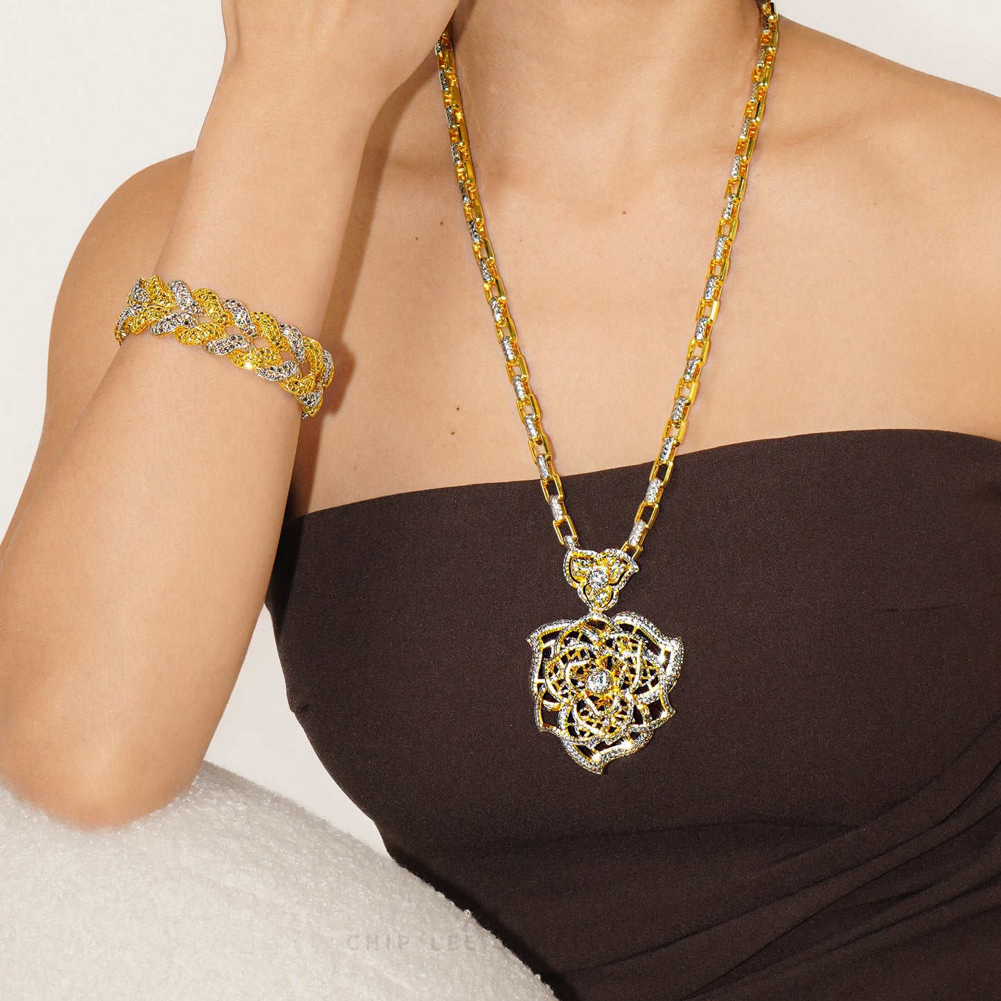 Model wearing a gold and silver-toned link necklace with a large openwork floral pendant and a chunky matching bracelet, styled with a dark strapless top.
