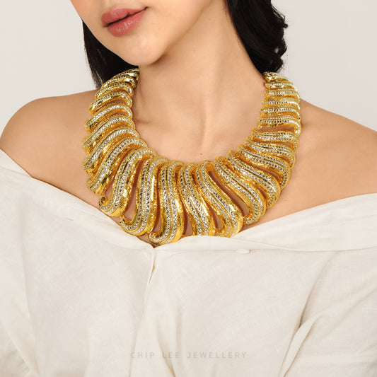 A close-up of a model wearing a chunky, wide gold statement necklace with textured, layered scales. The model is wearing an off-the-shoulder white top against a clean, light beige background.